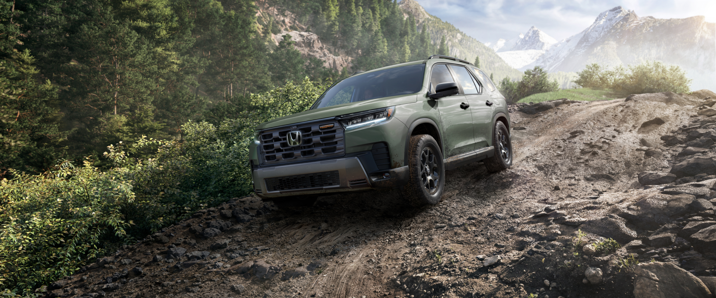 3/4 front side view of a green Pilot driving downhill on a dirt path in the forest, with distant mountains in the background.