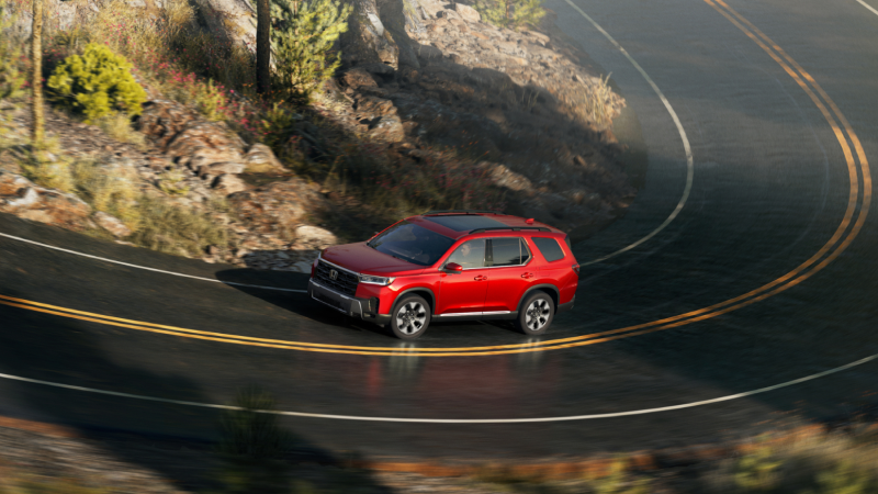 Side view of a red Pilot driving along a winding highway in a rugged landscape of rocks, bushes, and trees.