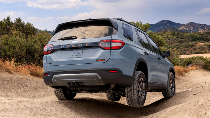 3/4 rear view of a light blue Pilot driving through uneven terrain with mountains in the background.