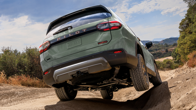 Rear view of a light green Pilot driving over uneven terrain with mountains and trees in the background.