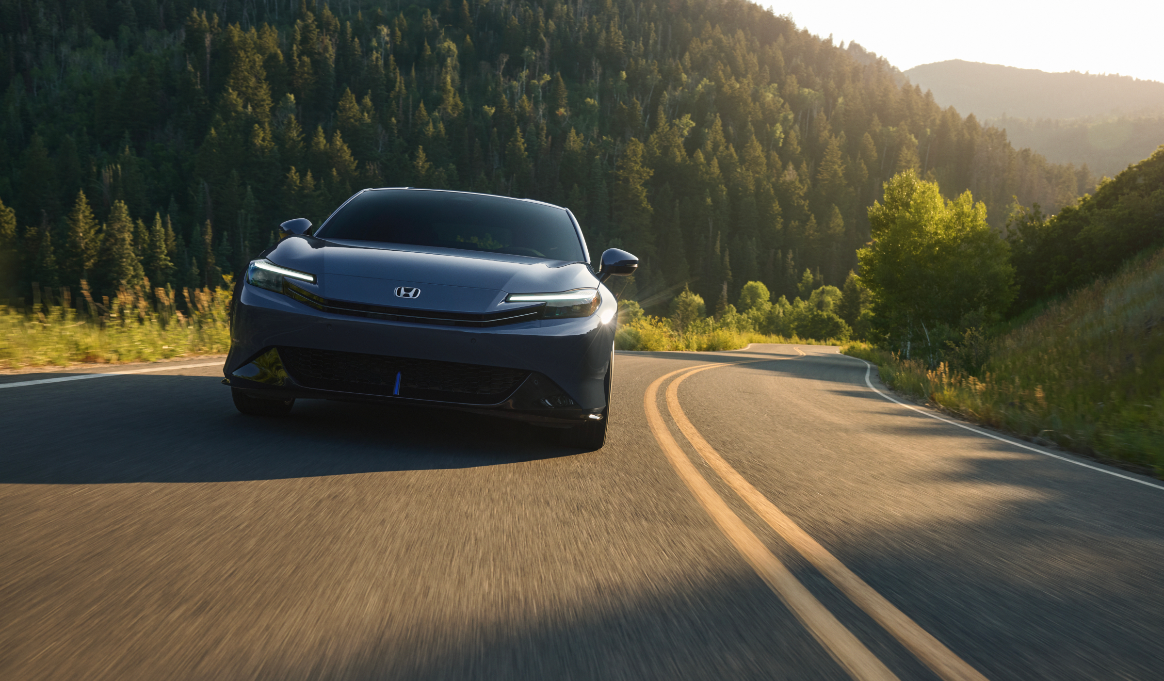 Front view of a Black Prelude driving on a highway framed by forest and warm light. 