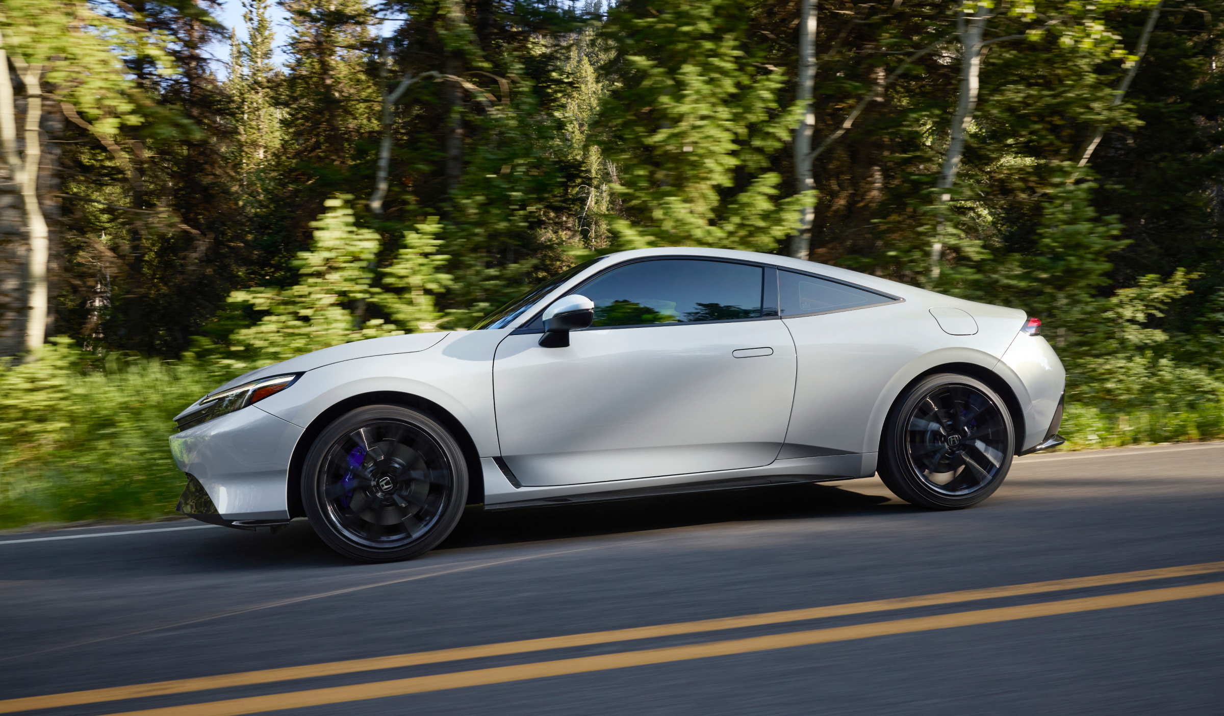 Side view of white Prelude driving on a road with forest in the background. 